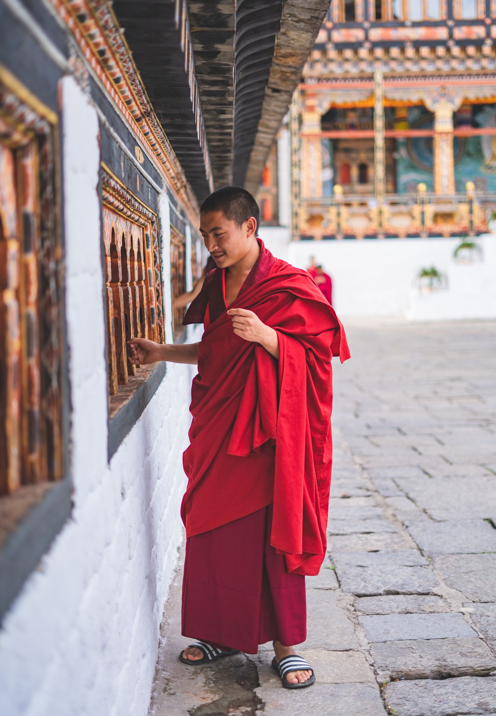 A young monk in red robes turning a row of prayer wheels along a whitewashed temple wall in Bhutan