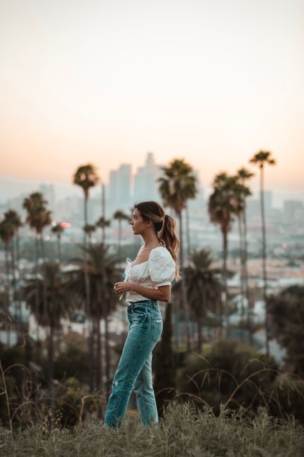 Woman in casual attire standing in front of Los Angeles skyline during sunset, surrounded by palm trees.