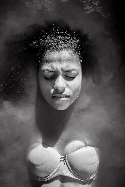 Black and white of African American female in swimwear lying in hot water under vapor with closed eyes