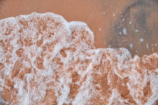 Foamy ocean waves gently washing onto sandy beach, showcasing natural texture and patterns.