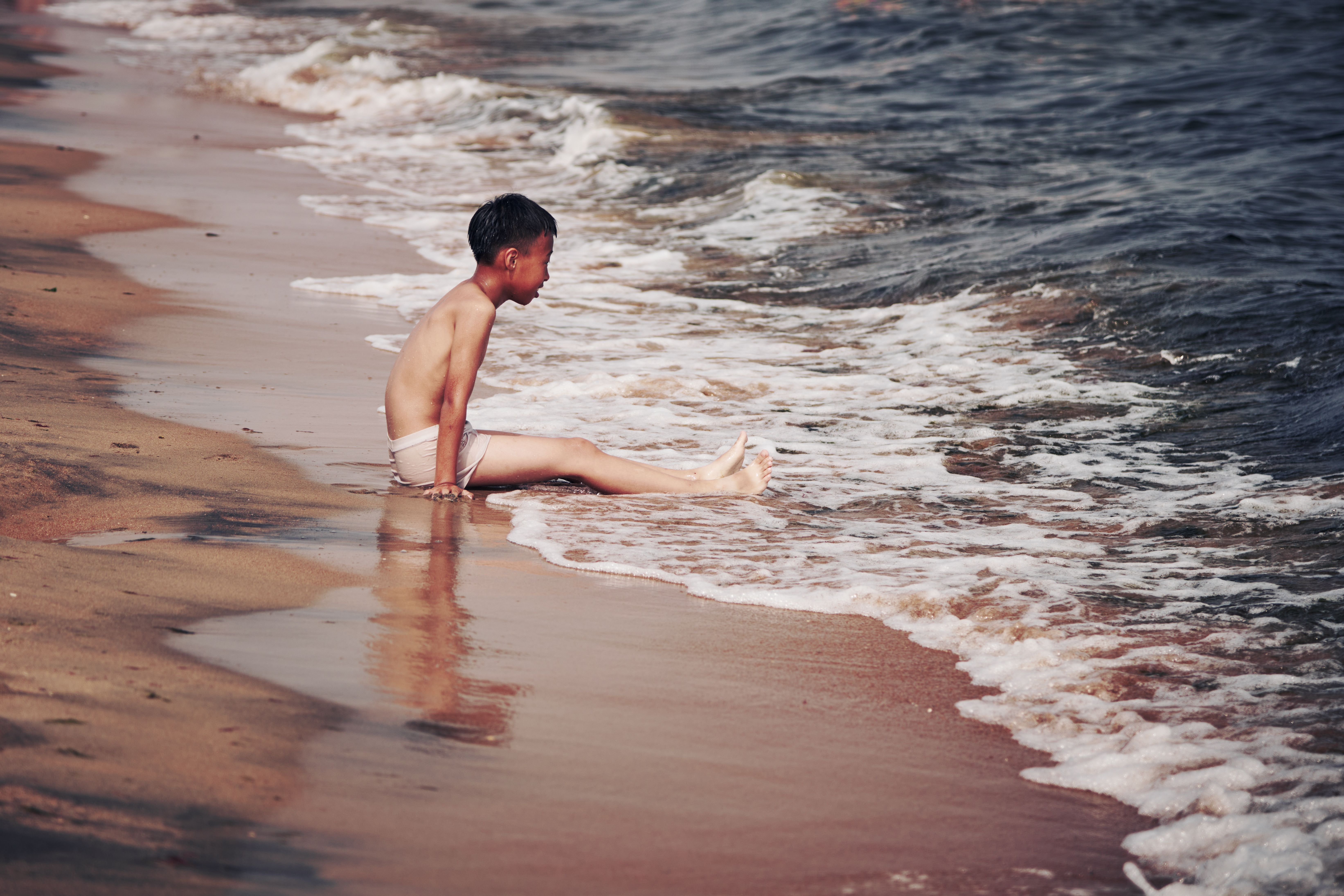 A child sitting by the seaside, interacting with waves on a sunny beach day.