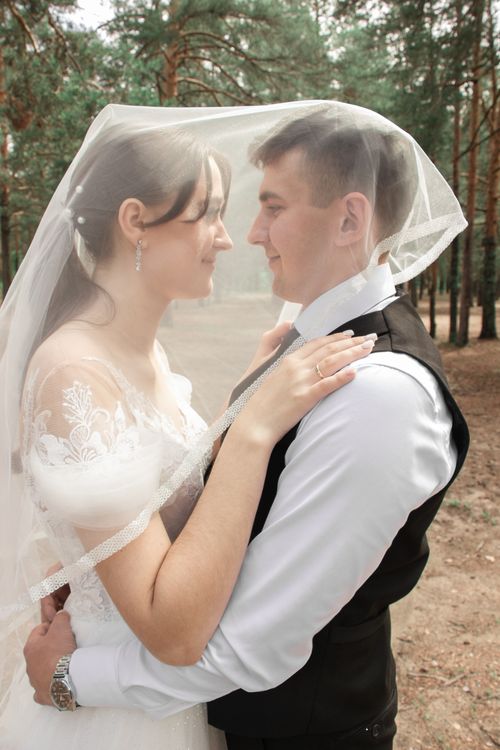 Free Elegant newlyweds embracing under a veil in a scenic forest setting. Stock Photo