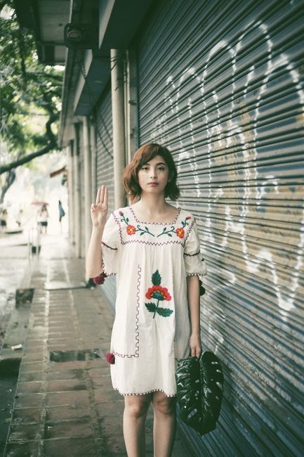 A young woman in a floral dress poses with a hand gesture near a roller shutter in an urban setting.