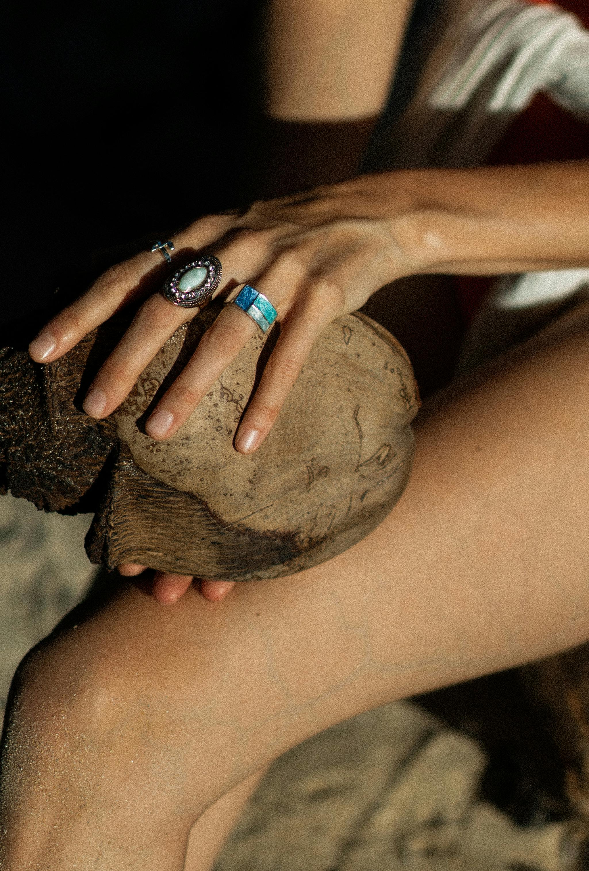Close-up of a woman's hand adorned with rings, holding a seashell on a sunny beach.