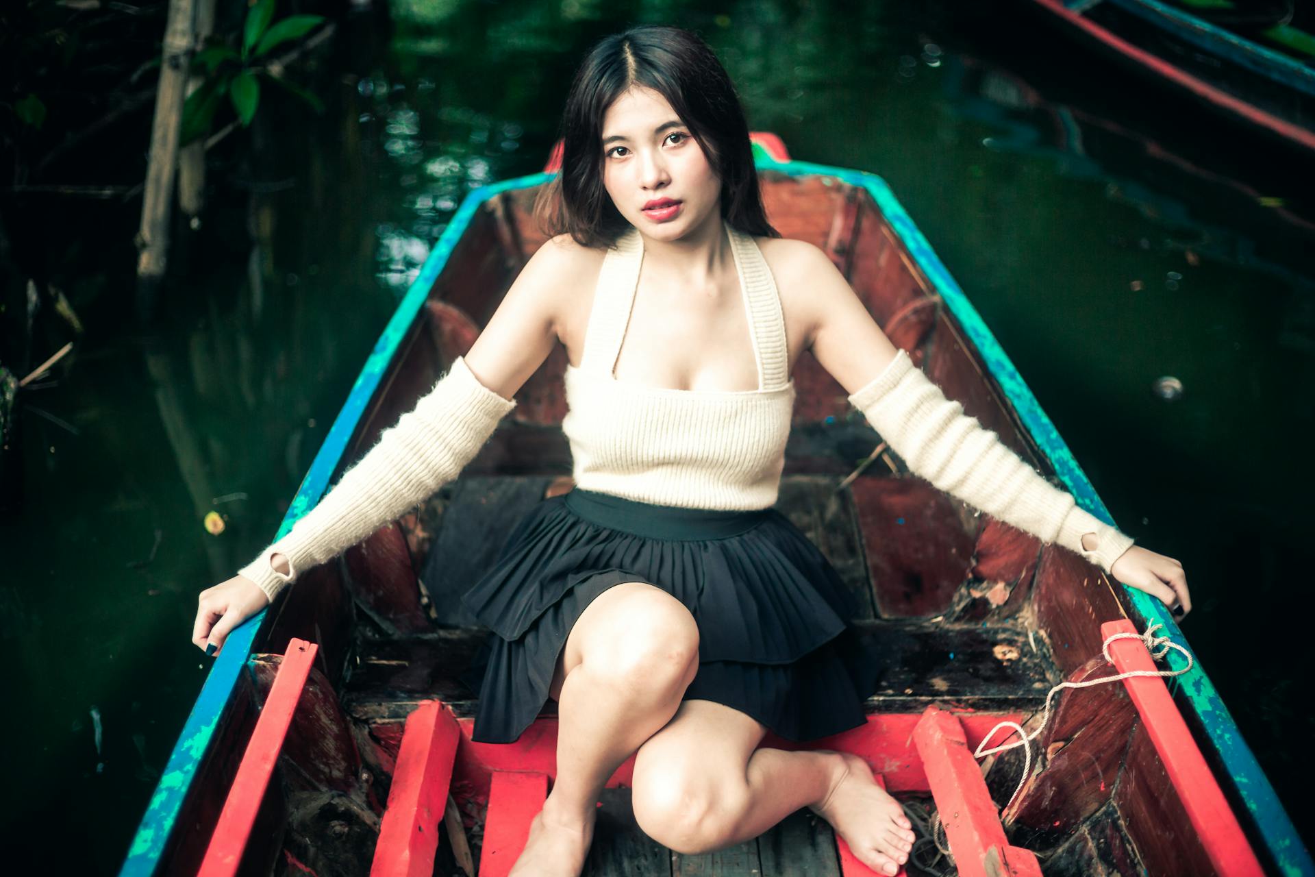 Asian woman poses elegantly in a boat wearing a stylish outfit, set against a serene river background.