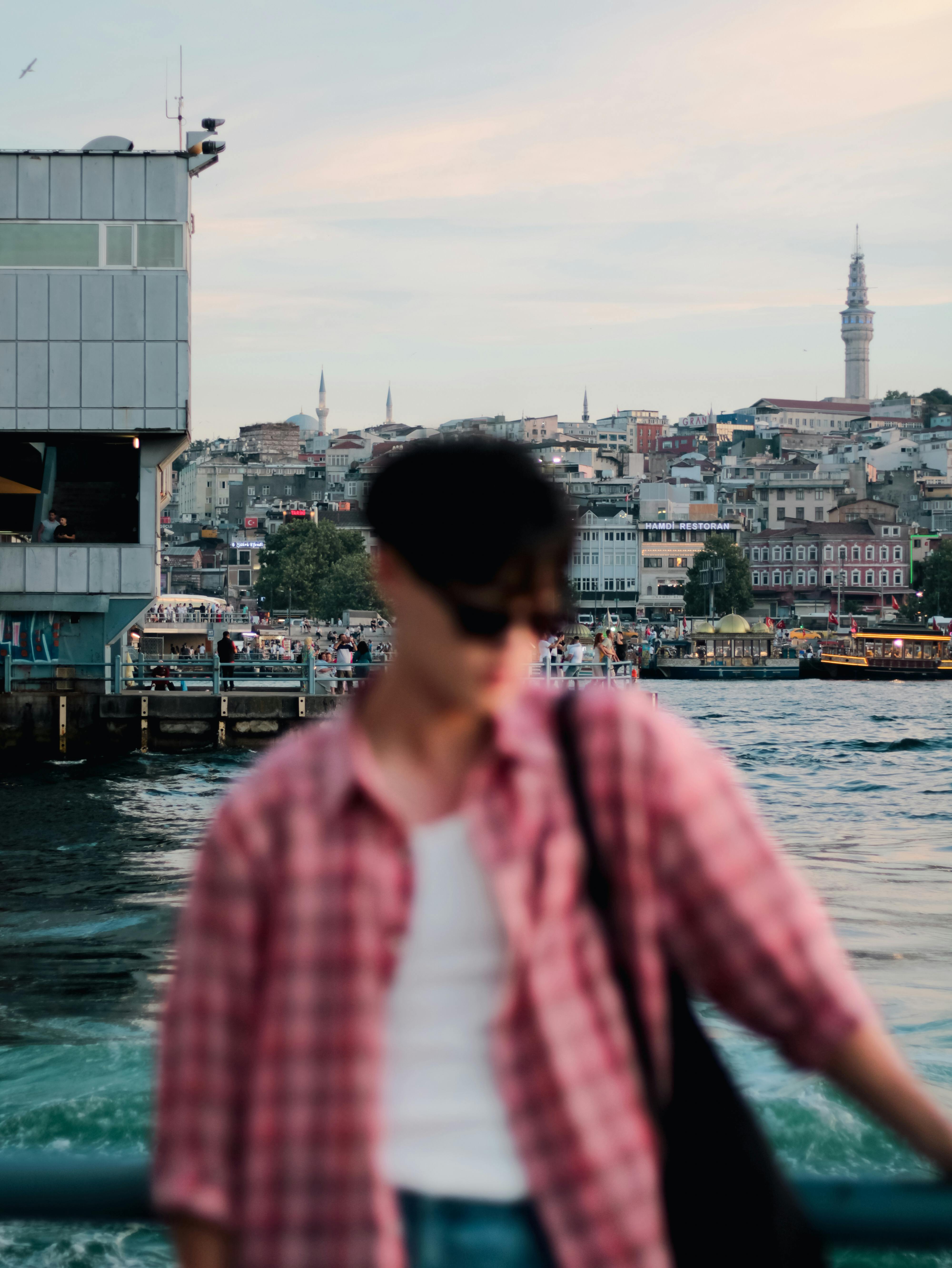 A stylish man stands by the seashore with Istanbul's cityscape in the background.