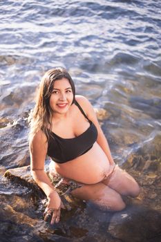 Pregnant woman in black top sitting in water, smiling on sunny day.