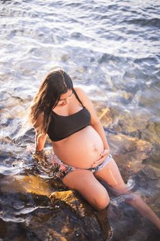 A serene image of a pregnant woman sitting on rocks by the sea, enjoying the warmth of summer.