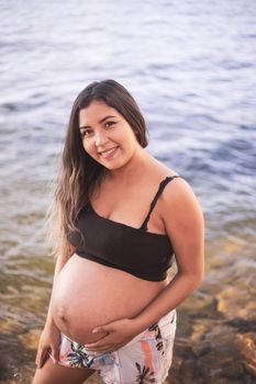 A happy pregnant woman with brown hair smiles by the seashore, capturing a serene outdoor moment.