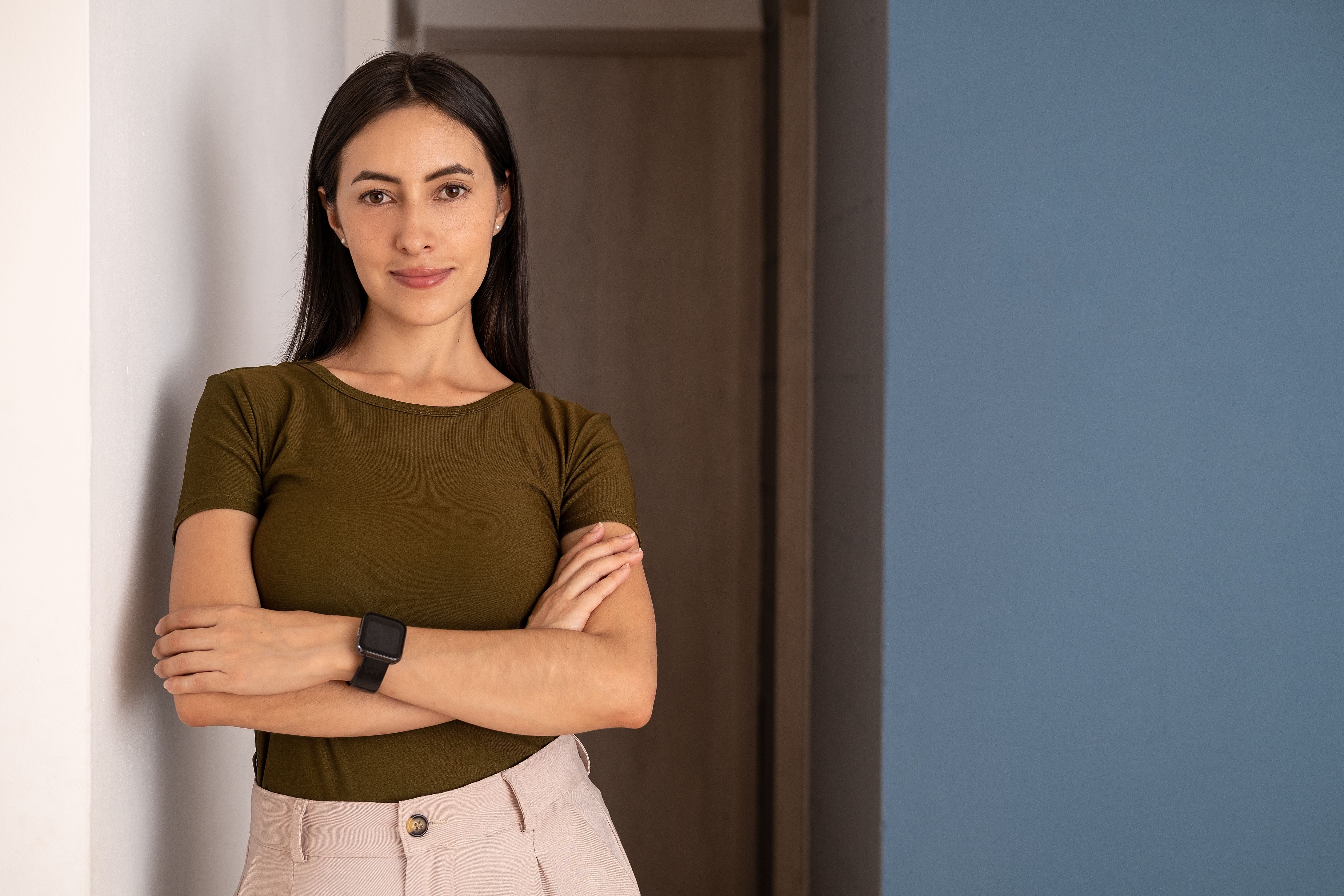 Portrait of a confident woman standing with arms crossed, wearing a smartwatch and casual attire indoors.