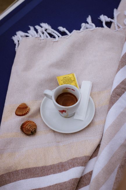 Espresso cup on beach towel next to seashells, perfect for beach vibes.