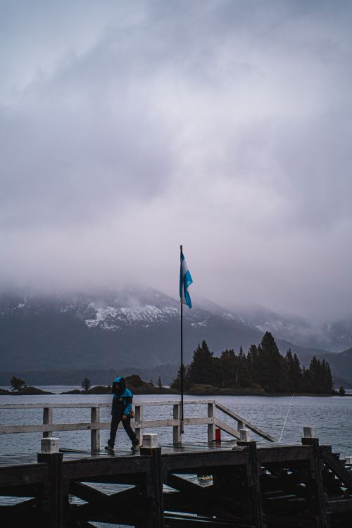 Free A lone figure walks on a pier in Bariloche, Argentina under a cloudy sky, with a flag waving. Stock Photo
