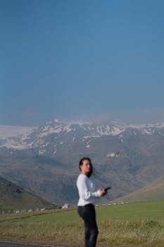 Woman controlling drone with snowcapped mountains in the background, clear sky above.