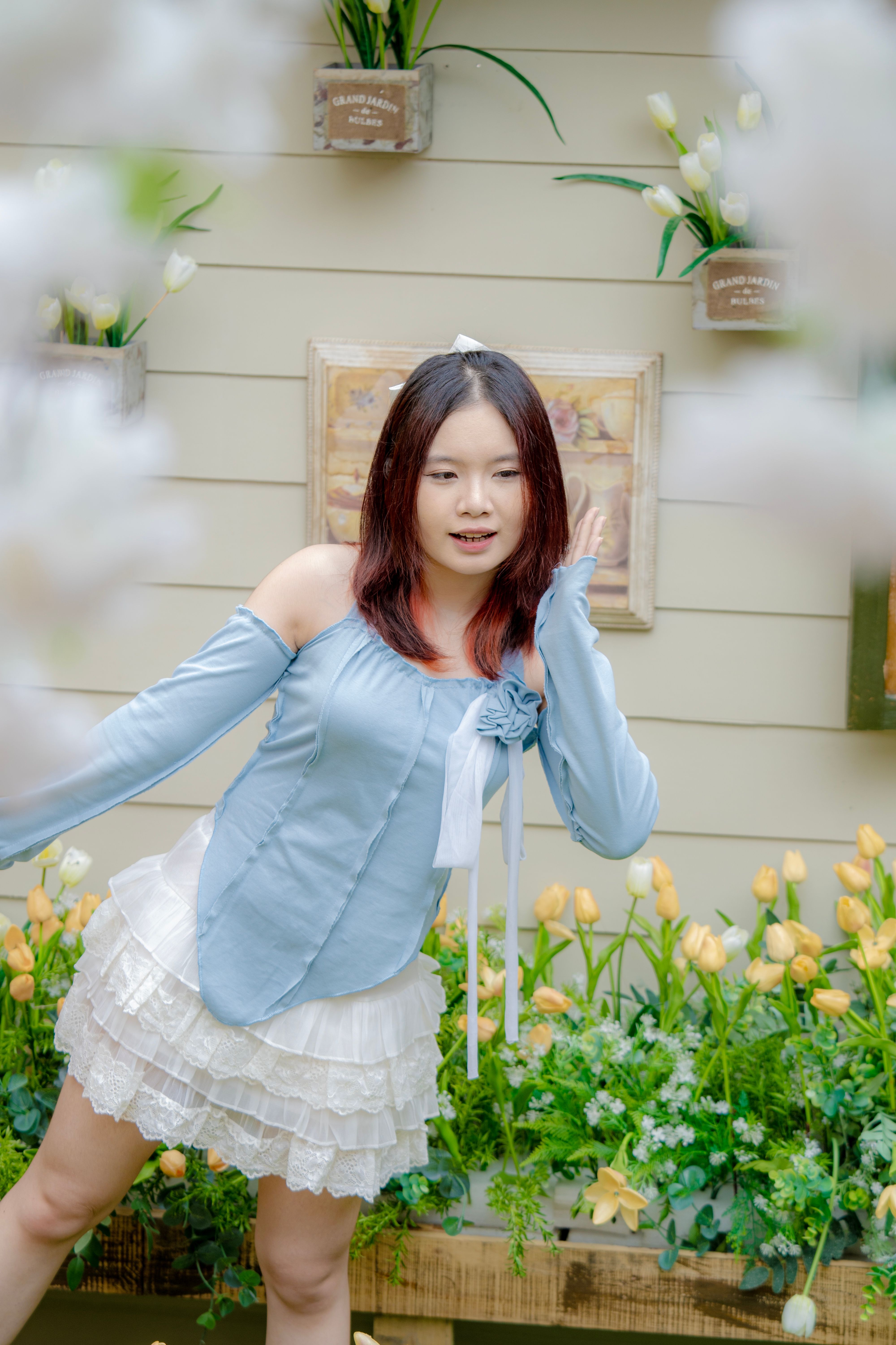 Asian woman posing in stylish blue blouse and layered skirt amidst garden flowers.