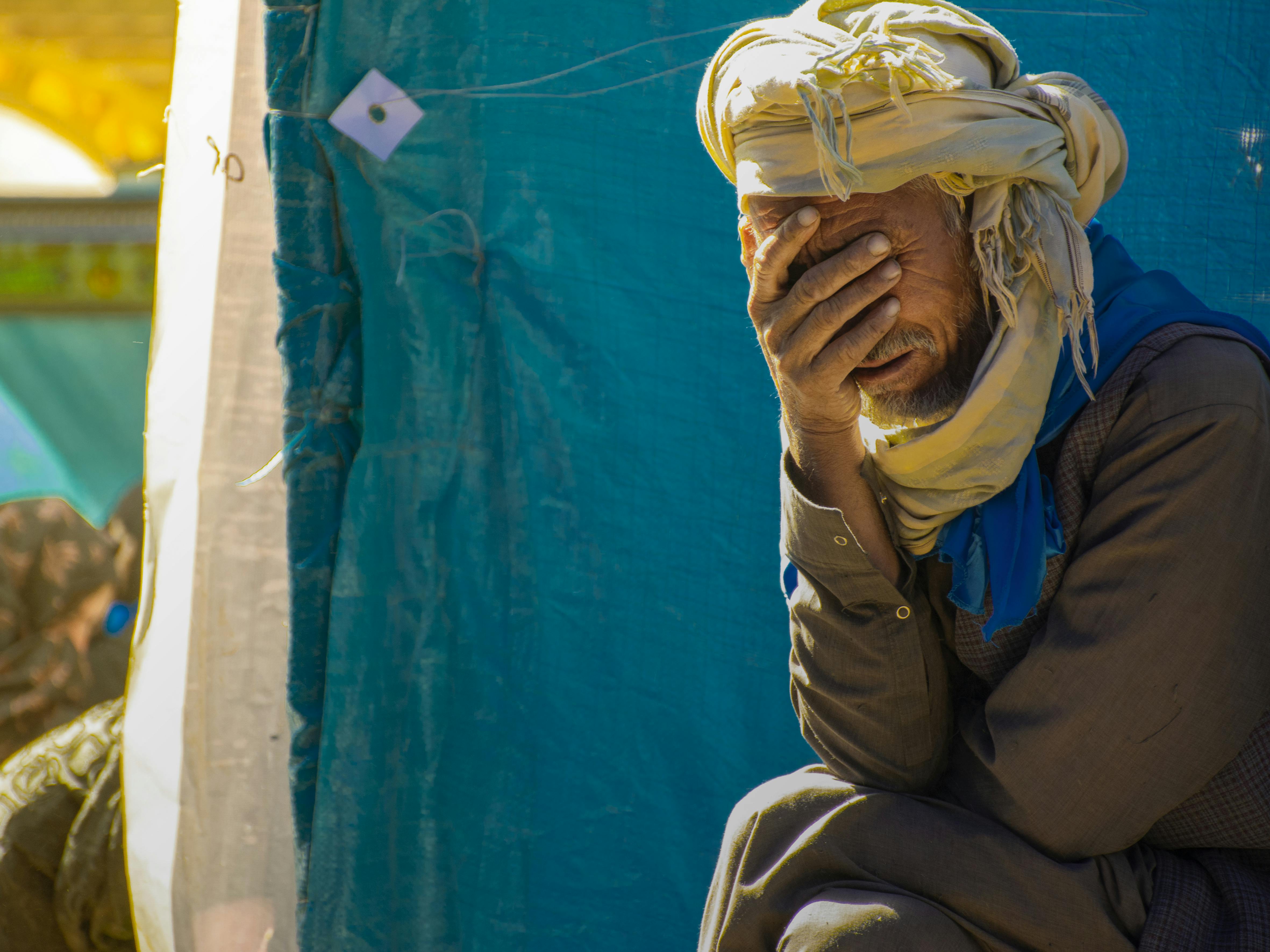 A senior man with a turban covers his face with his hand, expressing deep thought or emotion.
