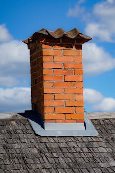 A classic brick chimney on a shingled rooftop with a clear blue sky background.