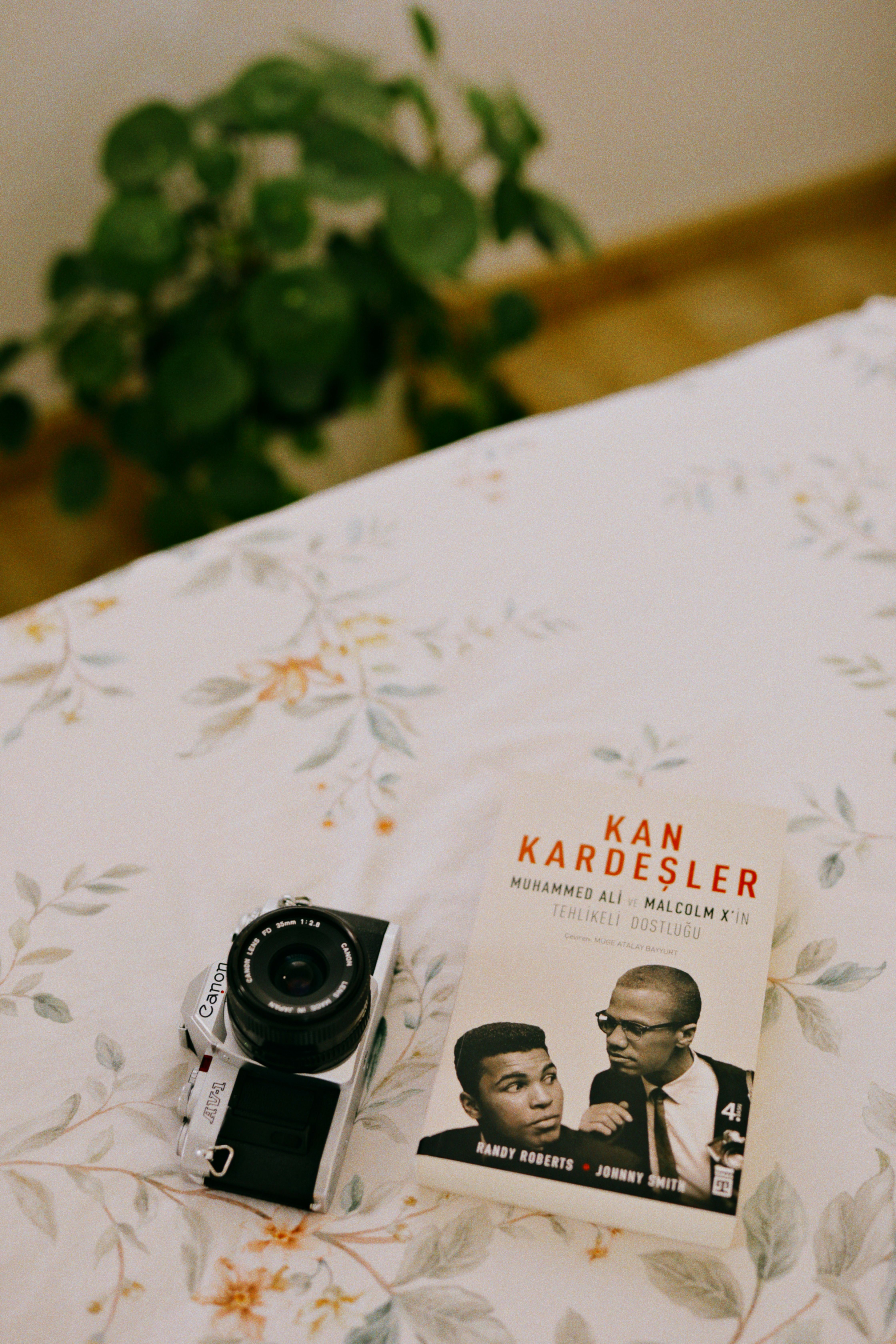 A vintage camera and book about Muhammad Ali and Malcolm X on a floral tablecloth.