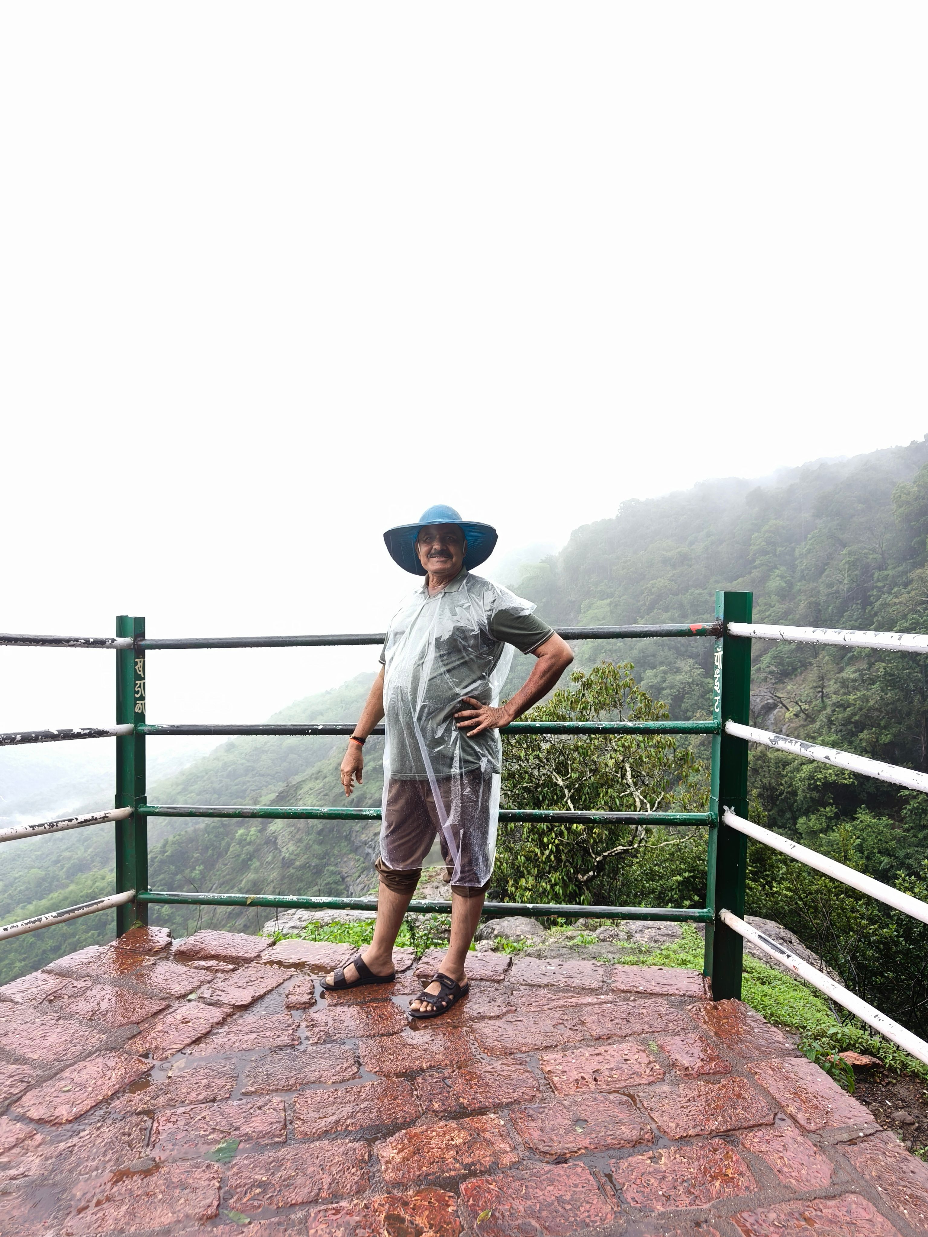 Senior man in blue hat stands on a terrace, overlooking foggy mountains with railing.