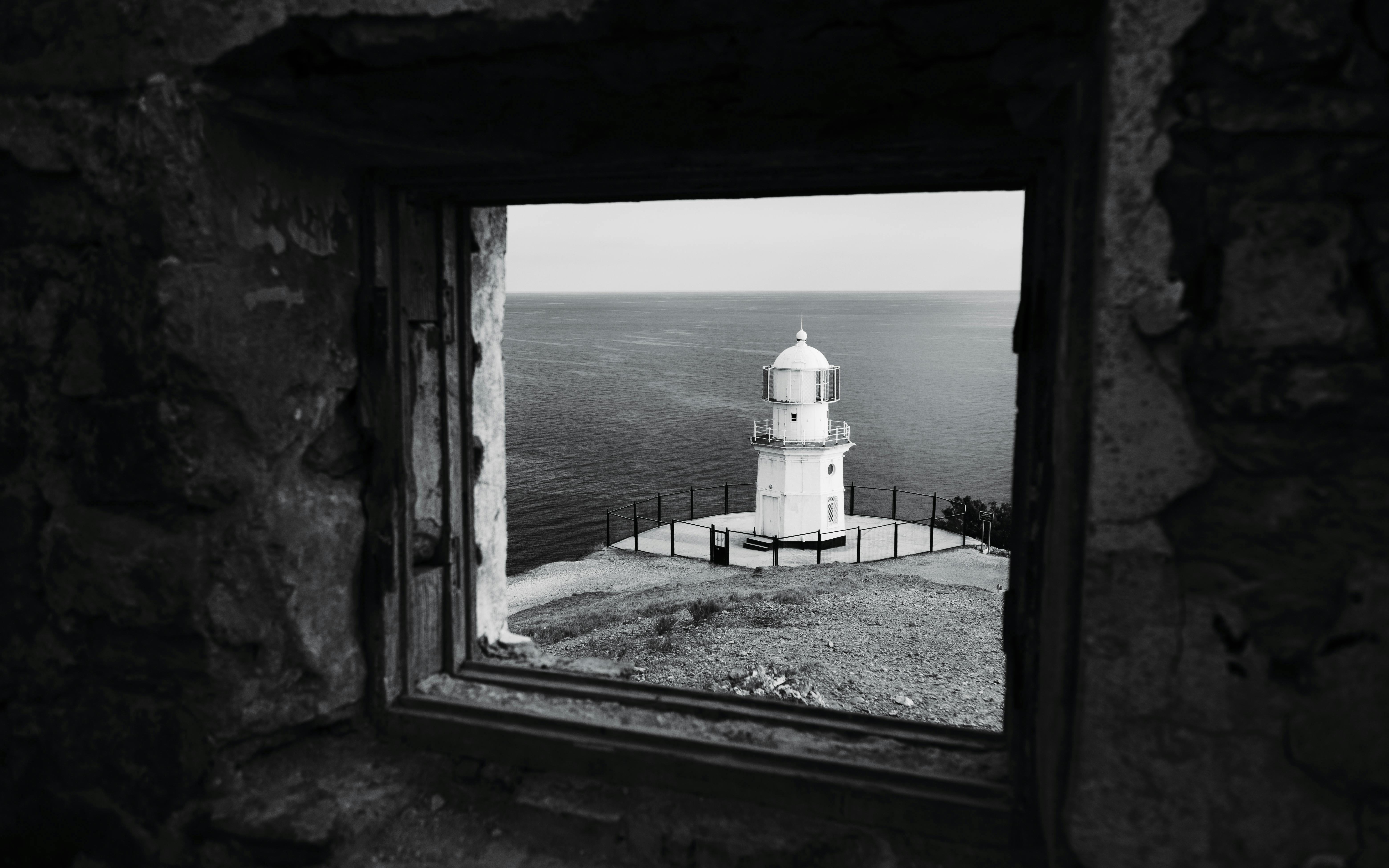 Black and white photo of a lighthouse viewed through an ancient stone window by the sea.