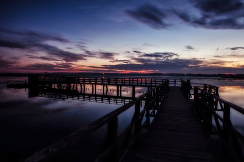 Free A tranquil sunset over a wooden pier, reflecting vibrant colors on calm waters. Stock Photo