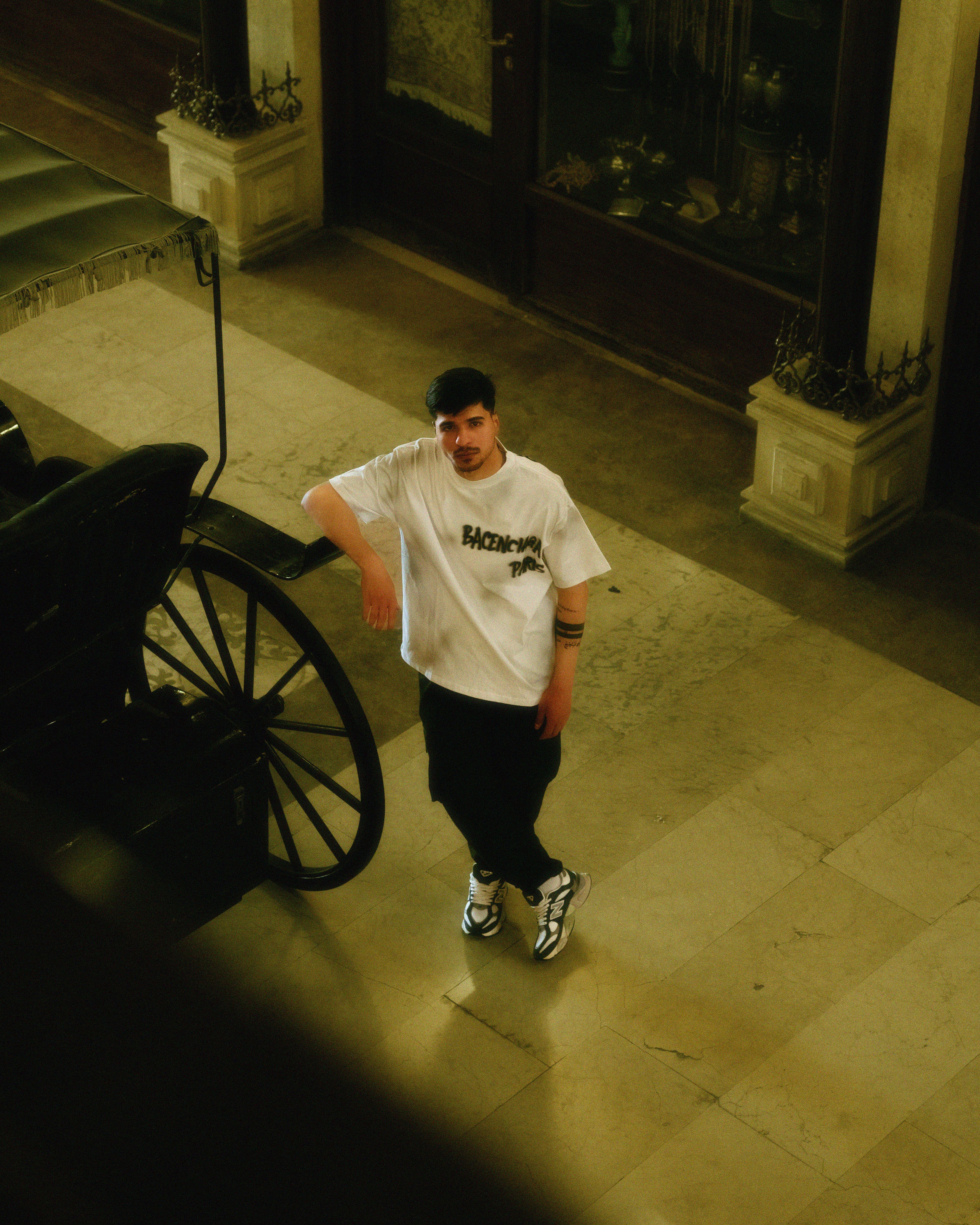 Trendy young man in stylish attire poses indoors with classic carriage backdrop.
