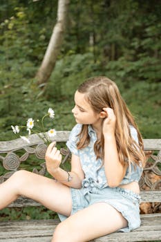 A young girl with long brown hair sits on a bench holding daisies, enjoying a peaceful outdoor moment.