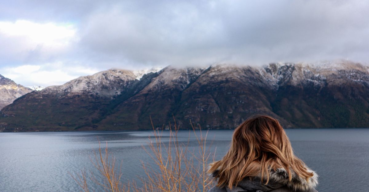 A woman with brown hair in a jacket gazes at Lake Wakatipu and snow-capped mountains in Queenstown, New Zealand.