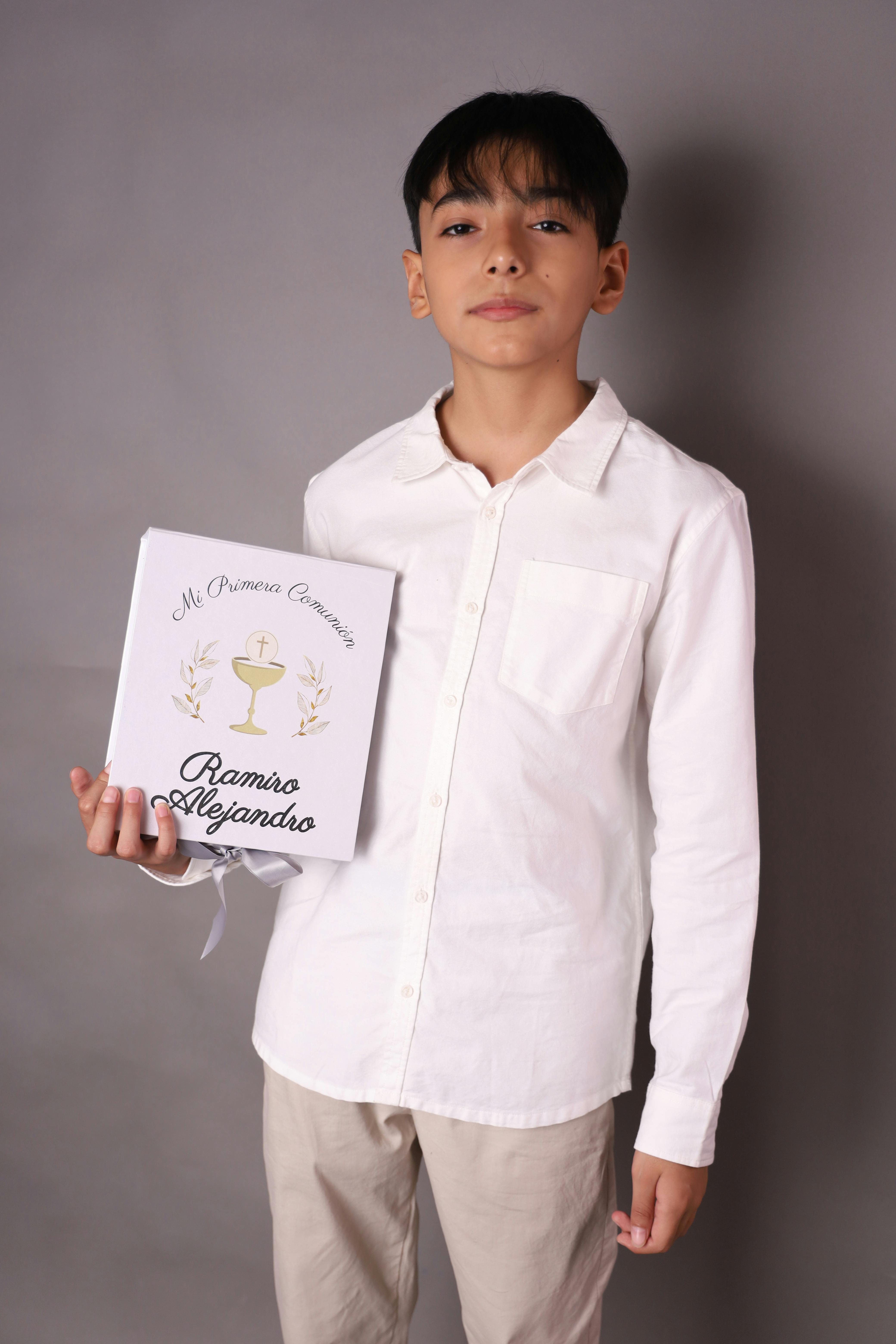 Boy with white shirt holding First Communion souvenir in a studio setting.