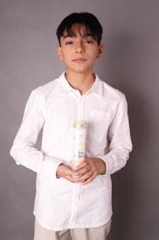 A young boy in a white shirt holding a bottle against a gray background in a studio setting.