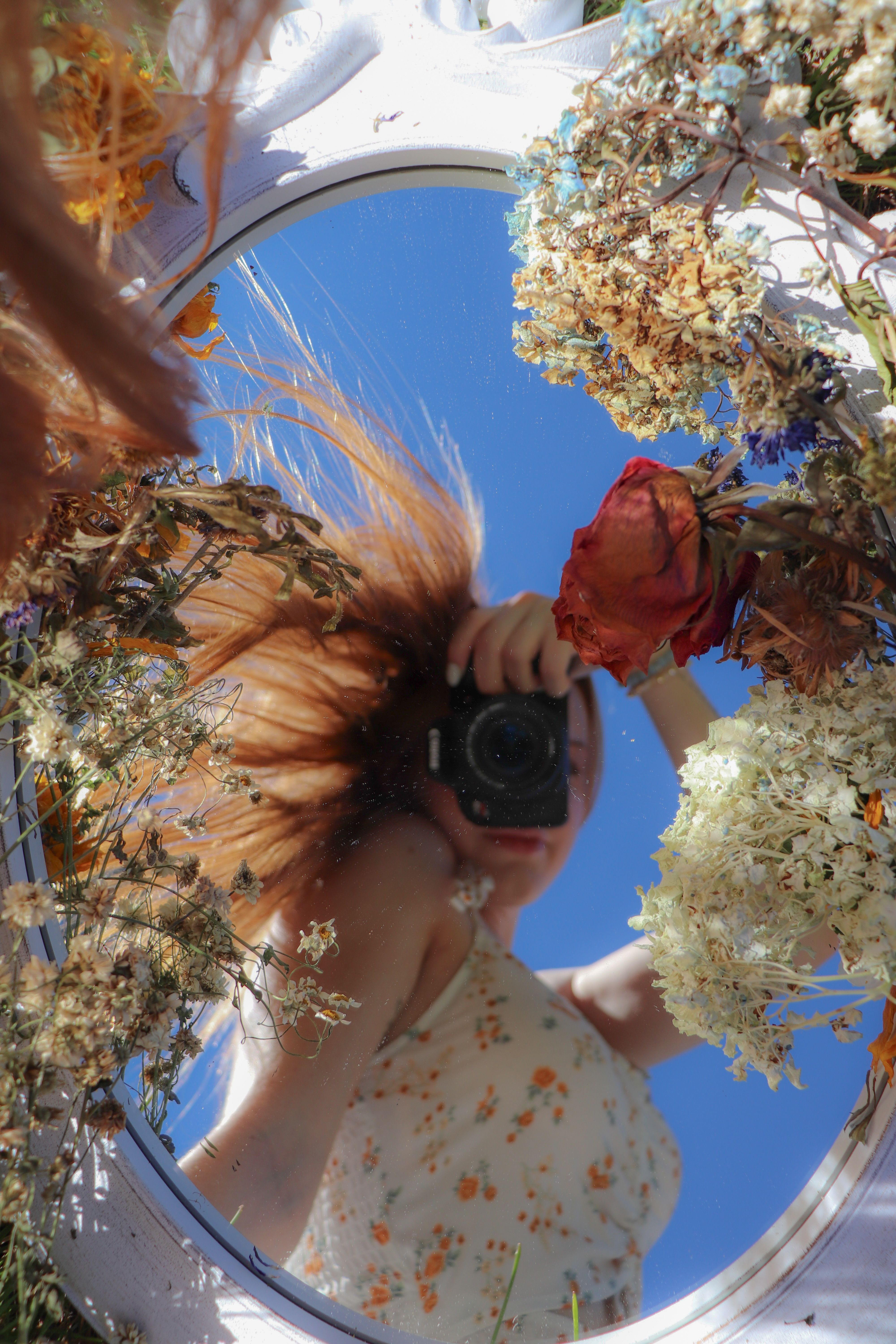 Artistic reflection of a woman in a mirror surrounded by flowers, capturing summer vibes.