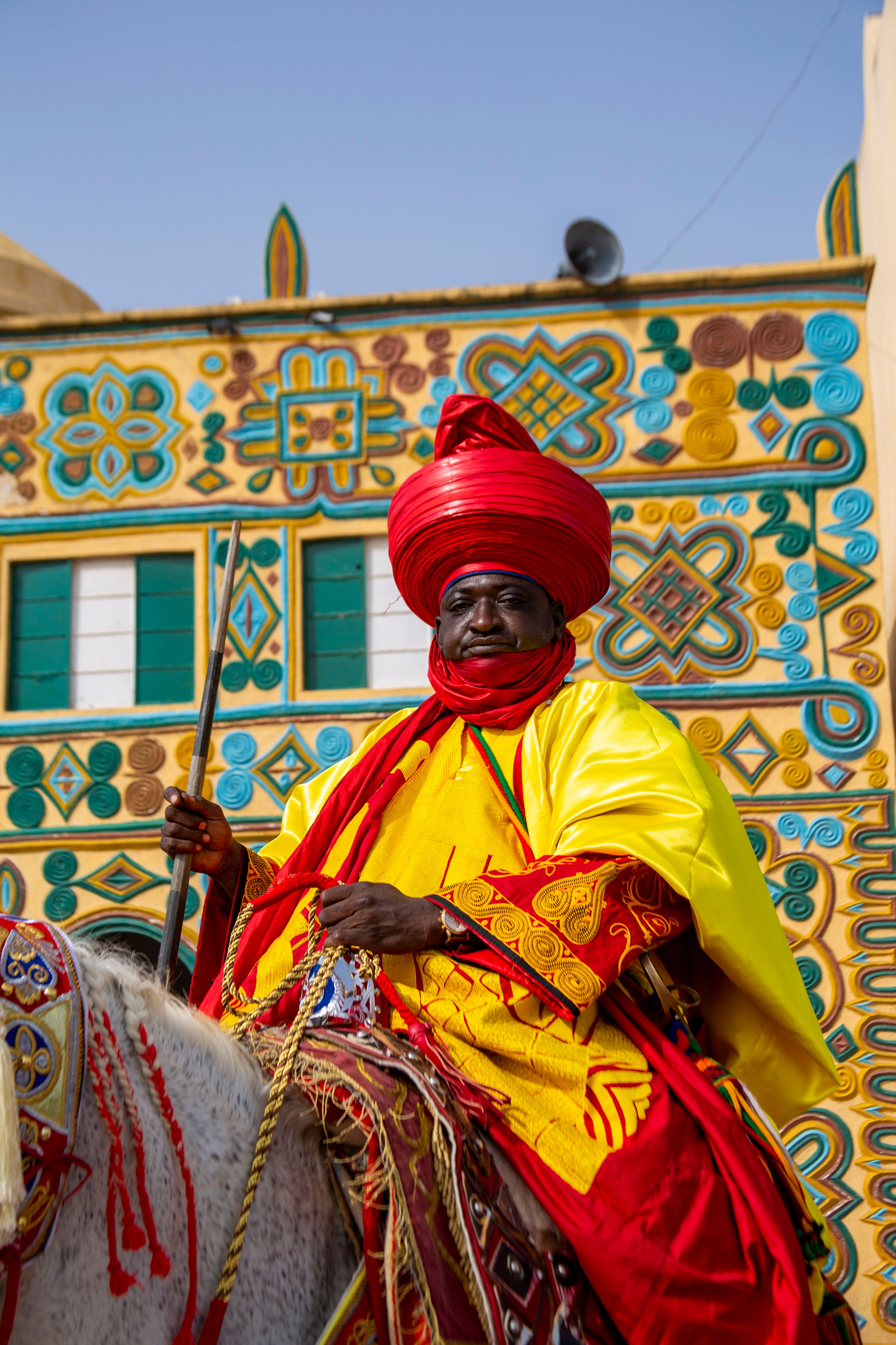Black man in traditional Nigerian attire horseback riding in front of an ornamental wall.