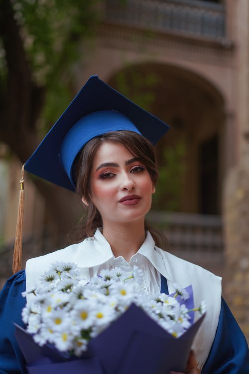 Free A young woman in academic dress and mortarboard holds flowers outdoors. Stock Photo