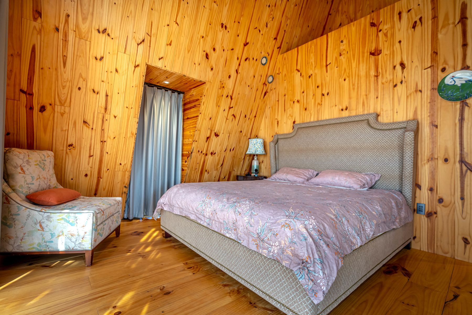 Wood-lined bedroom with an upholstered bed, floral armchair, patterned bedding and narrow curtained doorway