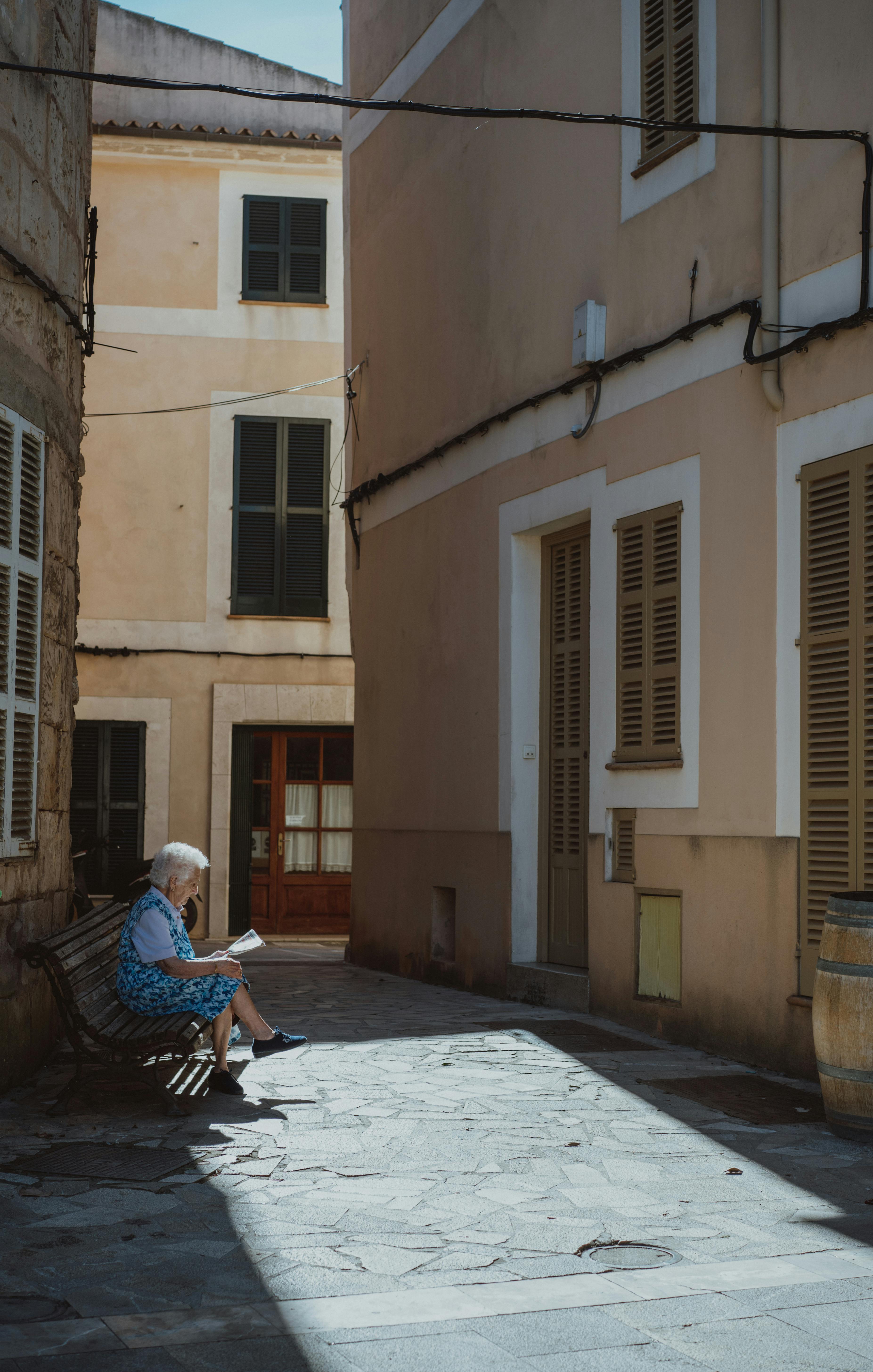 A serene scene of an elderly woman reading in a sunlit alleyway in Sineu, Spain.