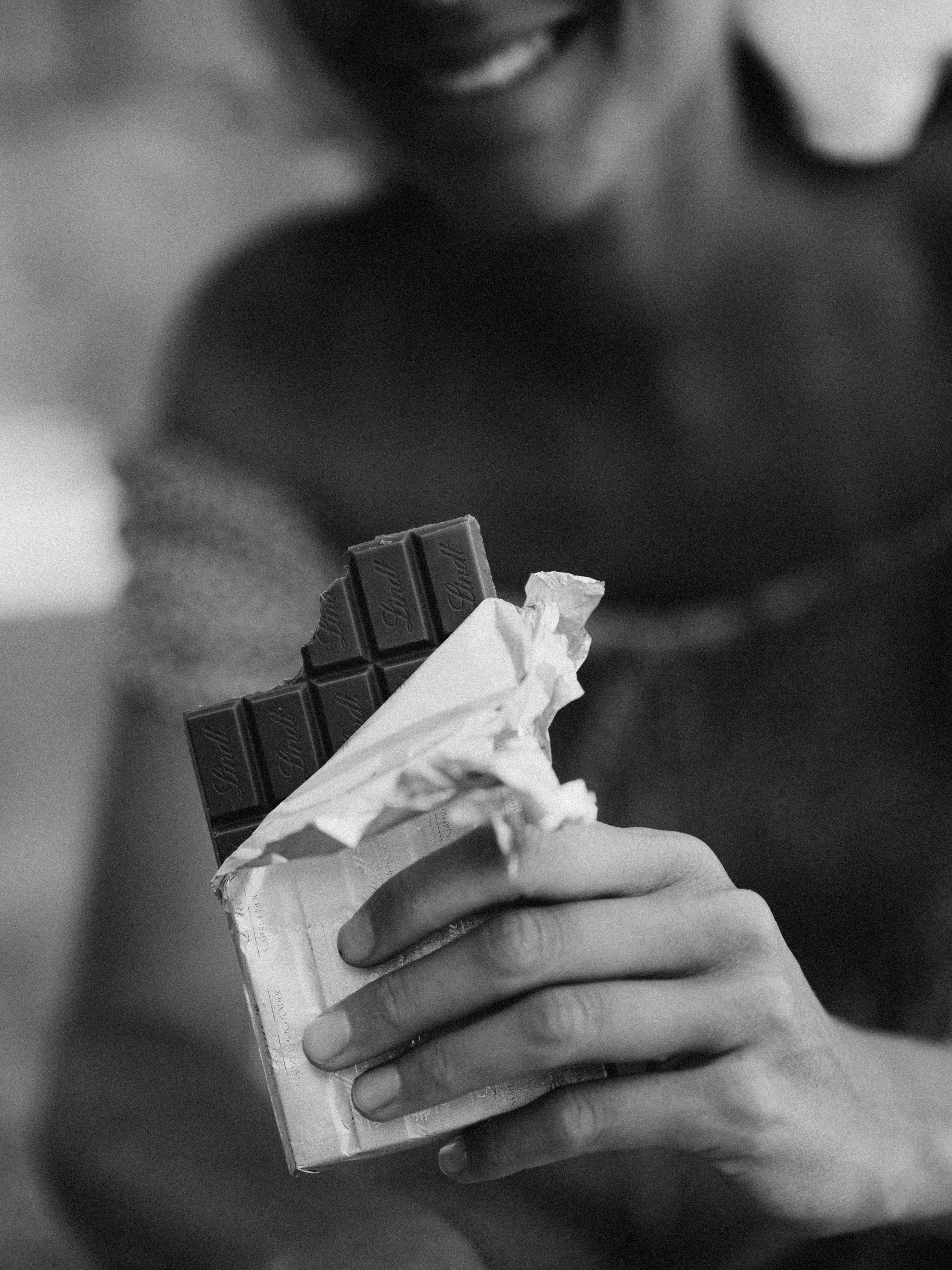 Monochrome image showing a hand holding an unwrapped chocolate bar with a smile in the background.