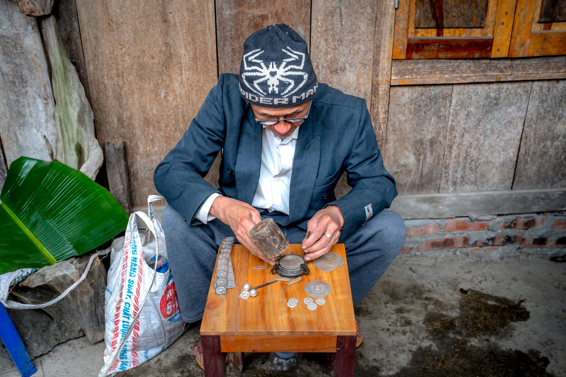 Senior man in winter cap working on metal items with coins on a wooden table in a rustic setting, focused on craftsmanship.