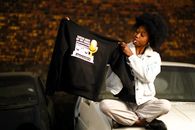 African woman sitting on car holding a t-shirt with hip hop radio awards design, cigarette in mouth.