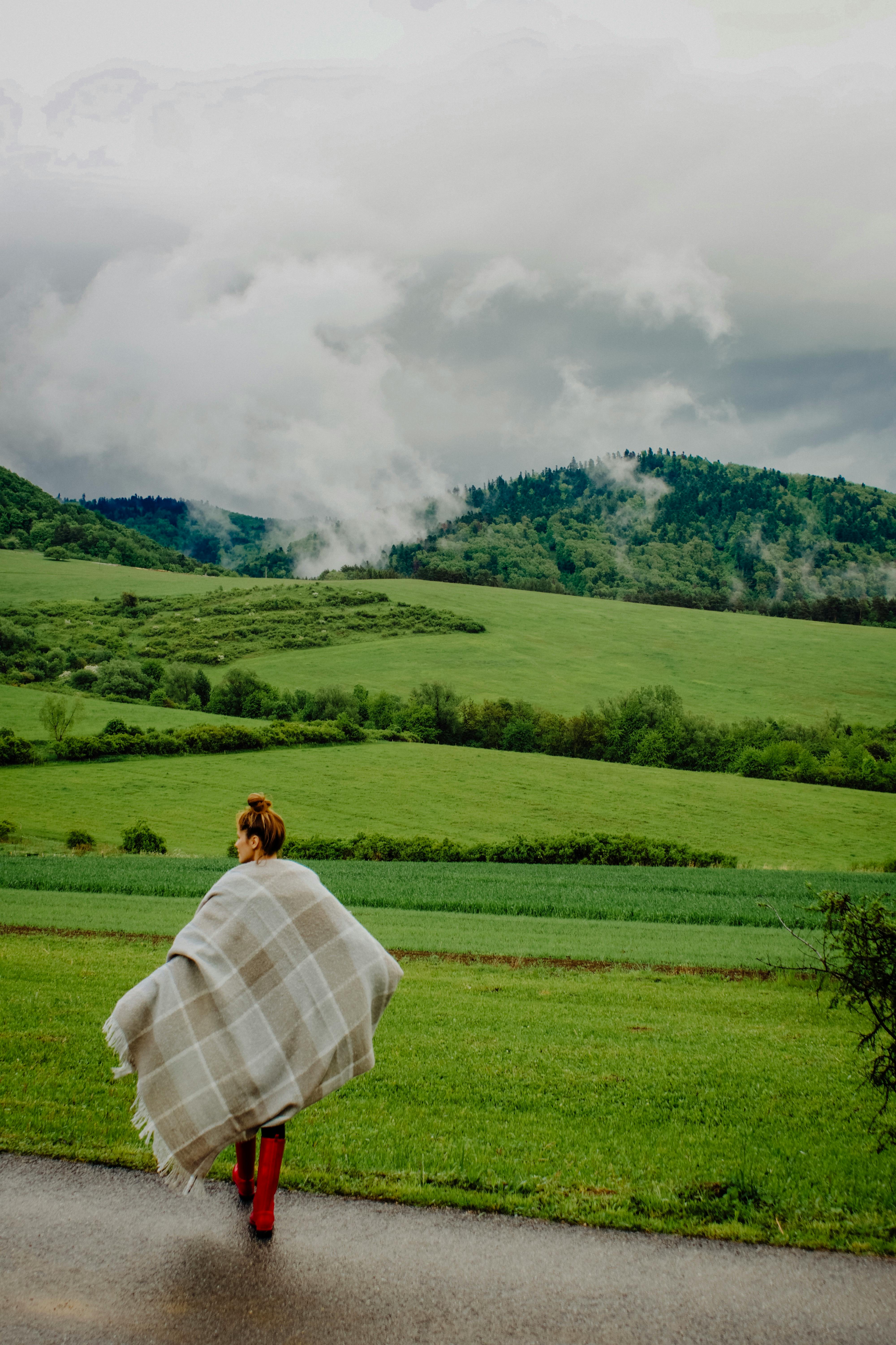 A woman stands with a blanket in a lush green landscape, capturing rural tranquility.