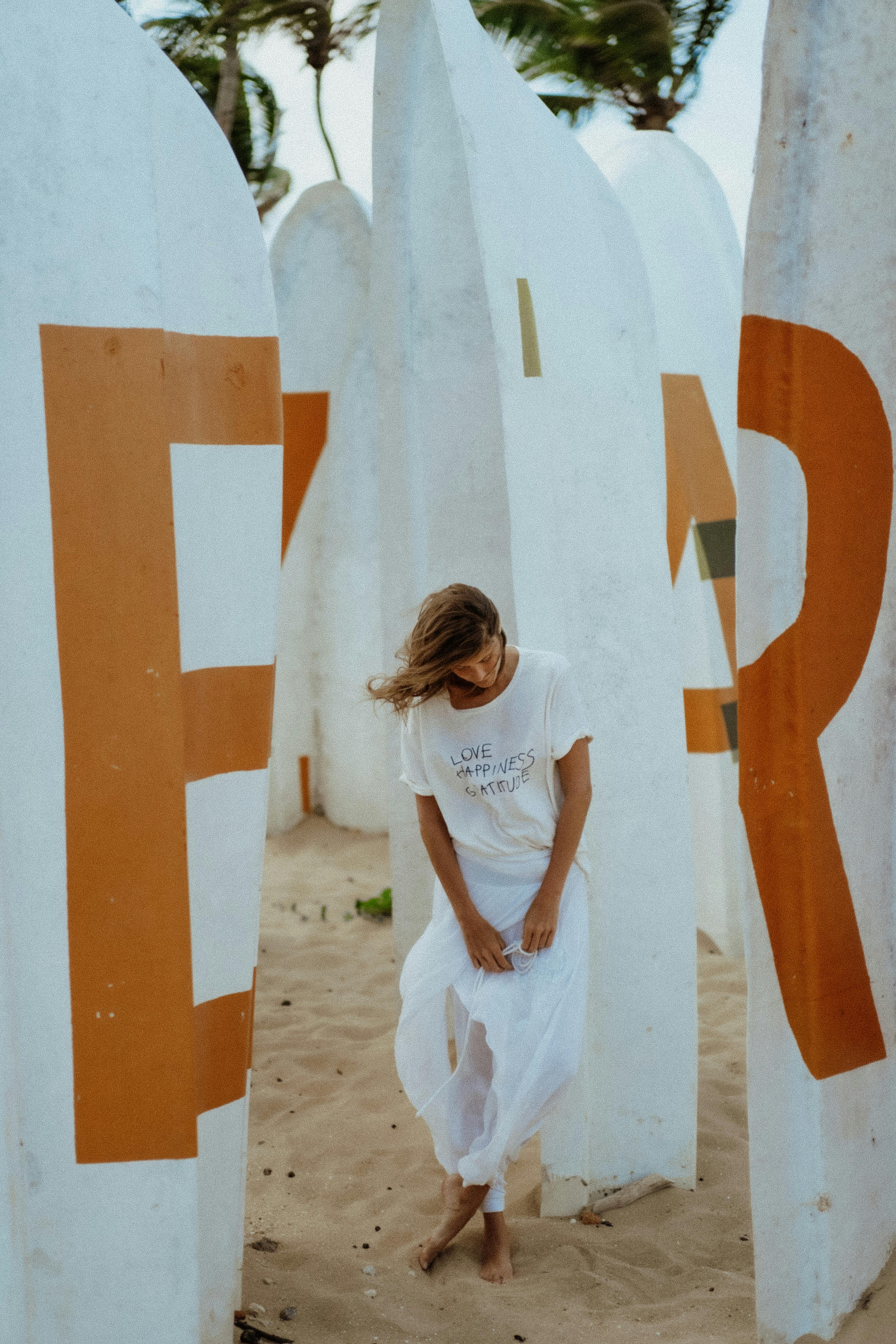 A woman in white clothing standing and posing between large artistic sculptures on a sandy beach.