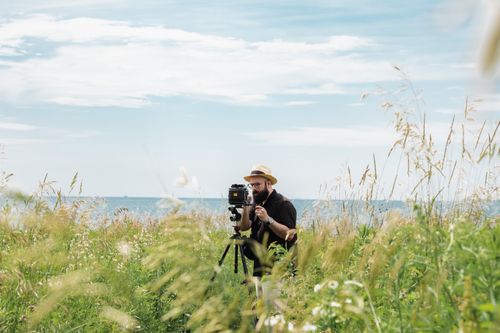 Free Man with camera and tripod takes photos in a scenic meadow overlooking water. Stock Photo
