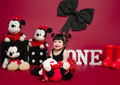 Charming baby girl in a Minnie Mouse themed setup with toys on red background.