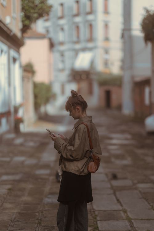 Free A woman stands in an urban alley, engrossed in her smartphone, capturing a candid moment. Stock Photo