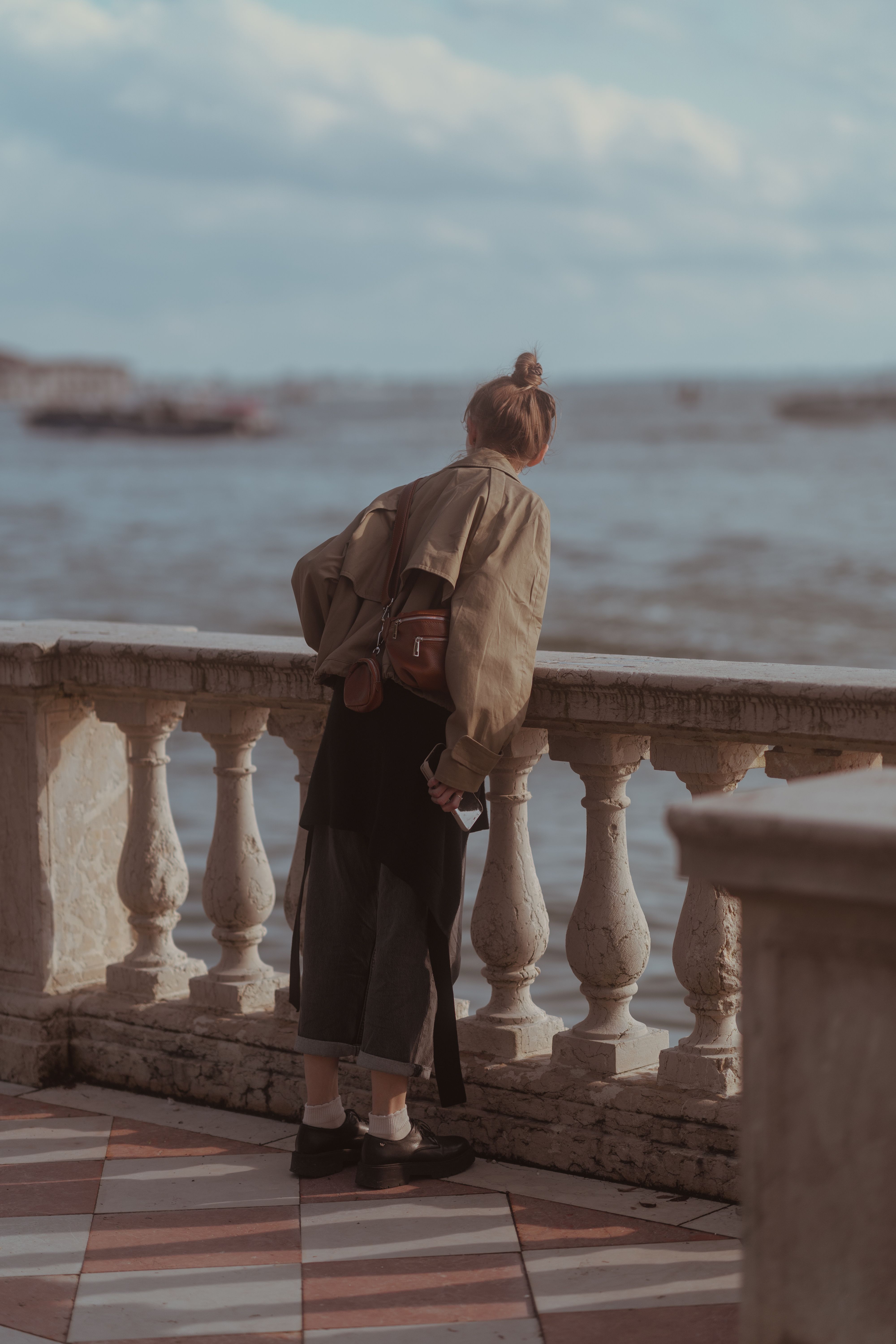 A woman leans on a balcony in Venice, Italy, overlooking a serene waterfront under a cloudy sky.