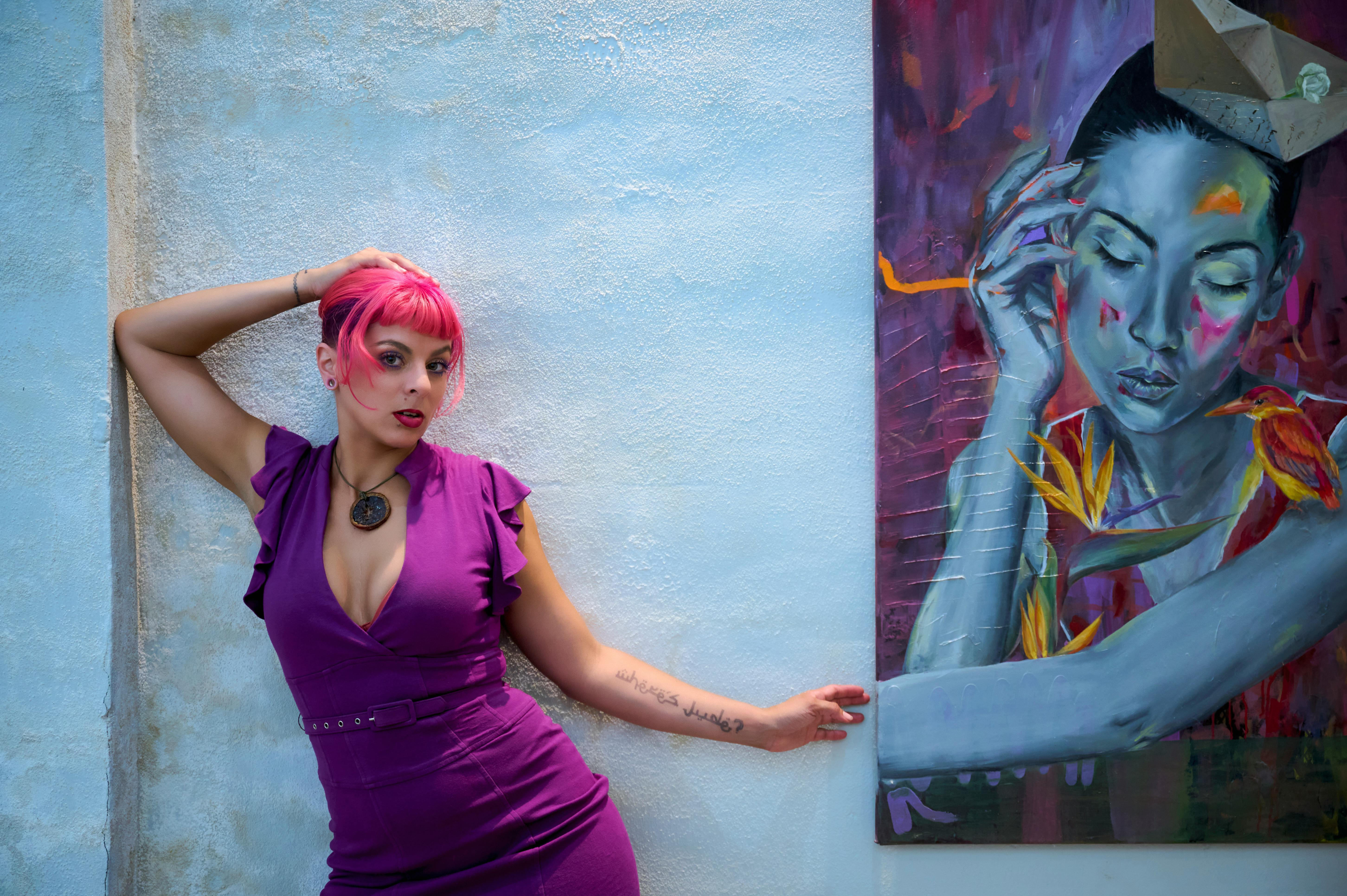 A stylish woman with pink hair posing against vibrant street art in a city setting.