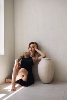 A beautiful woman in a black dress sitting by a large vase in a minimalist room.