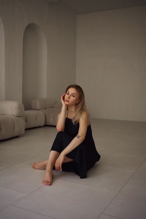 Free A beautiful woman in a black dress poses barefoot on a minimalist indoor floor. Stock Photo