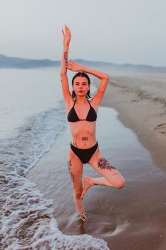A woman poses in a yoga stance on a sandy beach at sunset, showcasing tattoos.