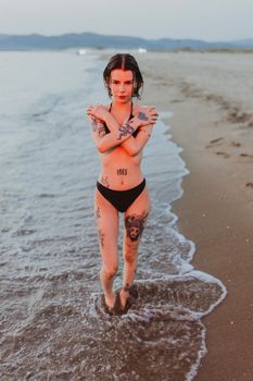 Stylish woman with tattoos standing on a serene beach during sunset.