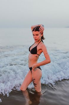 A tattooed woman poses confidently at the seashore in a bikini, surrounded by ocean waves.