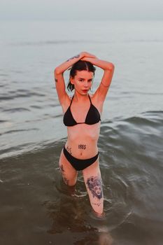 A woman with tattoos wearing a bikini poses confidently in the ocean at sunset.
